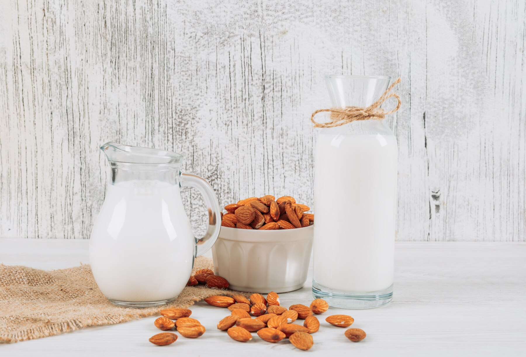side-view-milk-carafe-with-bowl-of-almonds-and-bottle-of-milk-on-white-wooden-and-piece-of-sack-background-horizontal_176474-4682.jpg